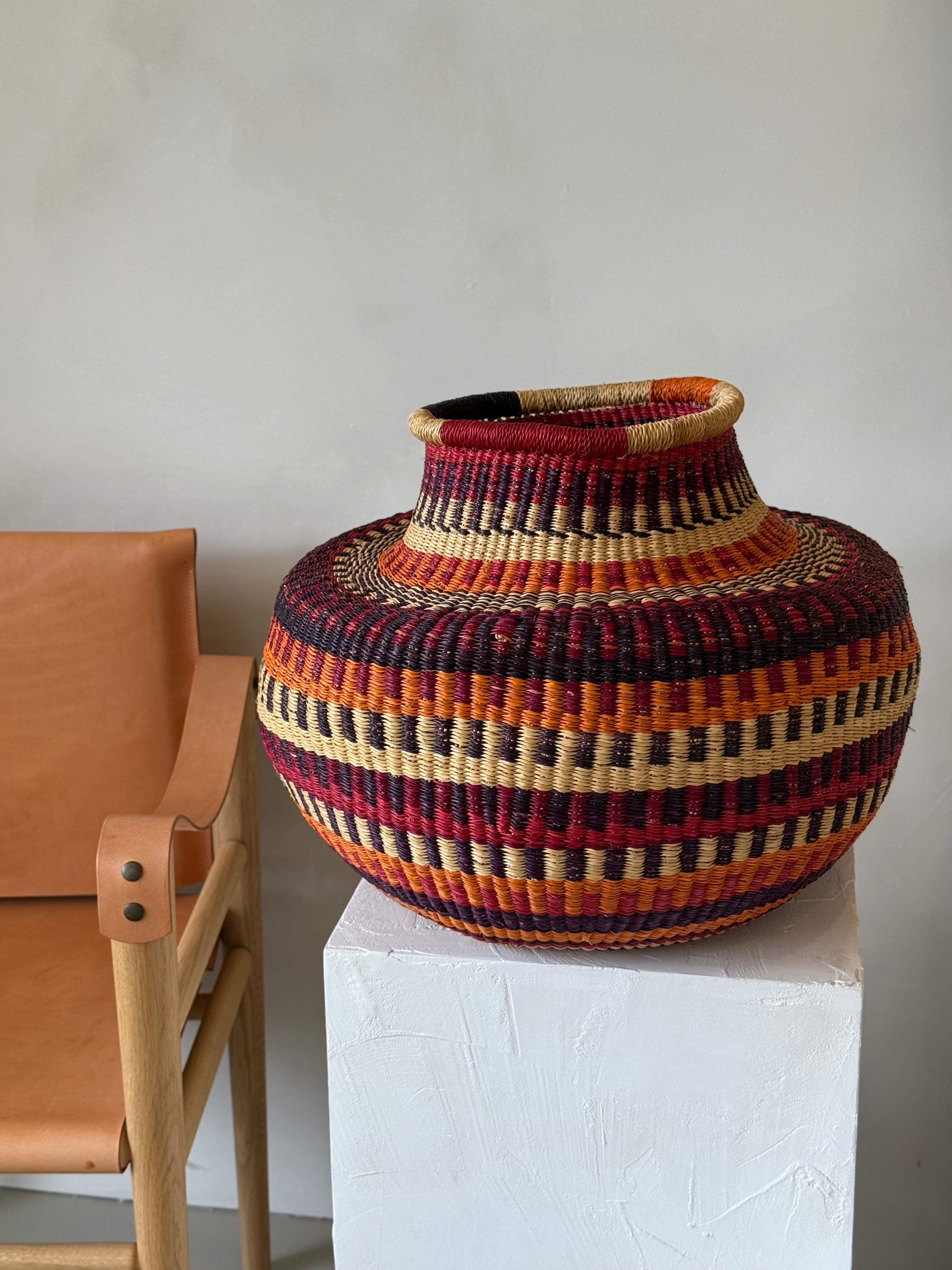 Large handwoven Ghanaian basket in red, amber, black and natural fibres beside a timber leather chair.