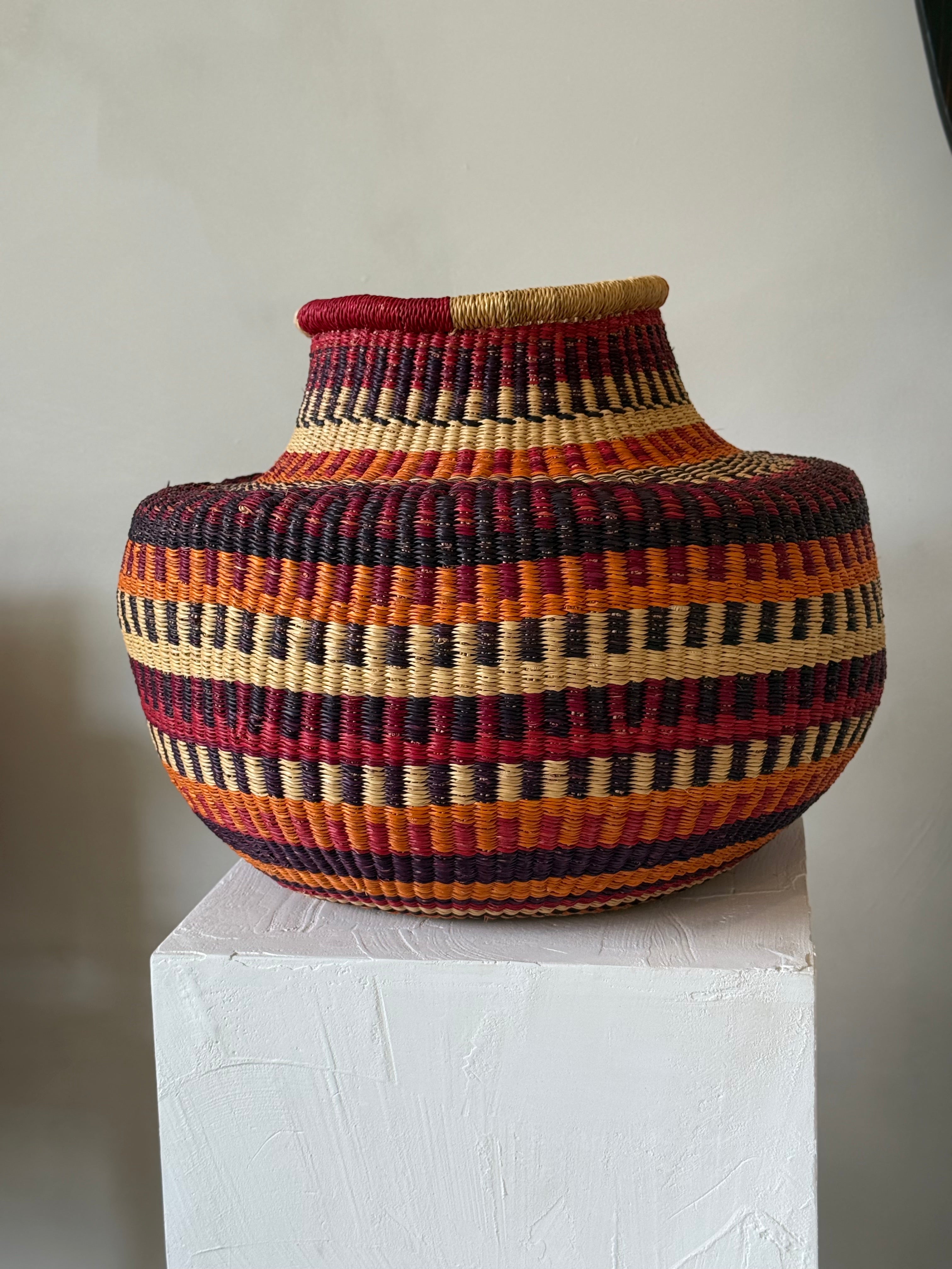Side view of red, orange and natural woven basket displayed on a white plinth.