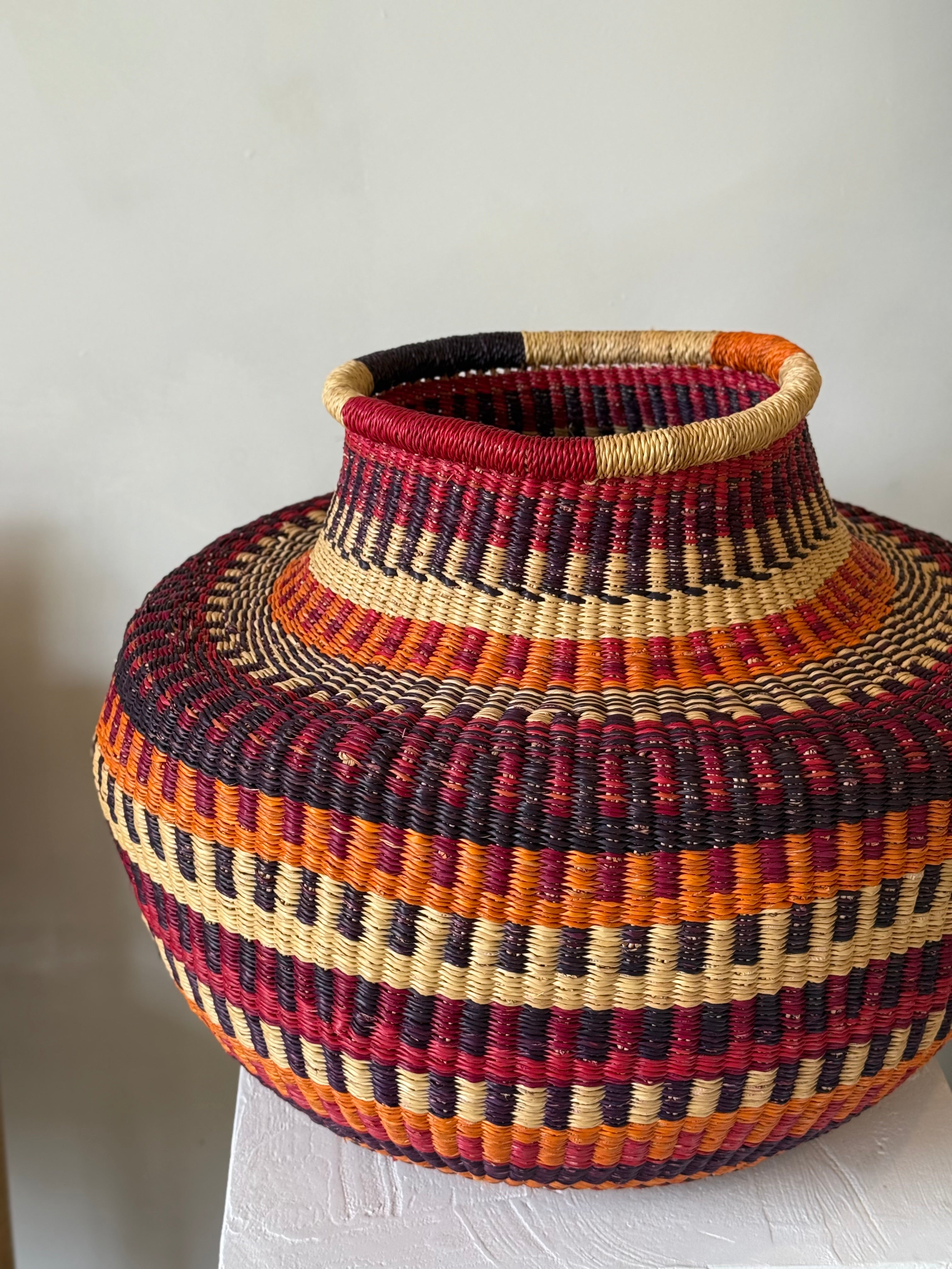 Sculptural Ghanaian basket in warm red and amber tones, showing tapered neck and rounded body.